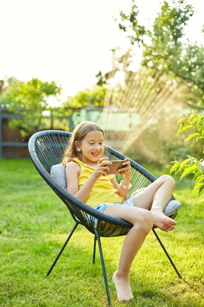 Happy kid girl playing game on mobile phone in the park outdoor, child using smartphone at home garden, backyard, sunlight, smartphone addiction