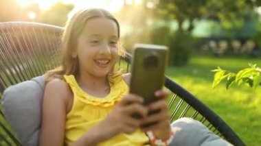 Happy kid girl holding smartphone having video call with friend distantly, online talk in the park outdoor, child using smartphone at home garden, backyard, sunlight, communication, technology concept