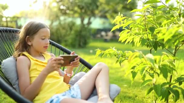 公園の屋外で携帯電話でゲームをプレイする幸せな子供の女の子 自宅の庭 スマートフォン中毒でスマートフォンを使用して子供 — ストック動画