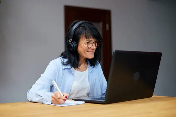 Smiling Mature woman wearing wireless headphones watch webinar, training, course, lessons on laptop take notes to paper notebook at home, smart senior female.