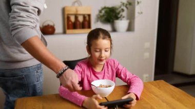 Little girl eating cereal with milk and play game on smartphone, dad takes the phone away, Slow motion of child having breakfast at home, dependence from gadget