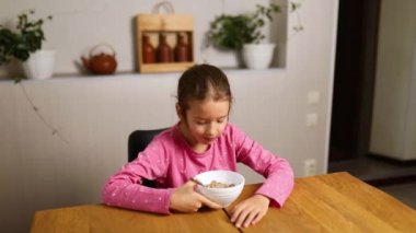 Little girl eating cereal with milk and take a phone to play game on smartphone, tasty healthy food, Slow motion of beautiful child having breakfast at home, dependence from gadget