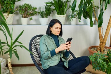 Woman chatting using smartphone, sitting on armchair at home, types message with happy smile, Concept of home garden, Biophilia design.