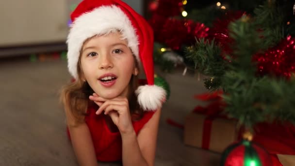 サンタ ハットの女の子が横になり 自宅でクリスマスツリーの近くで歌を歌う メリークリスマスとハッピーホリデー — ストック動画