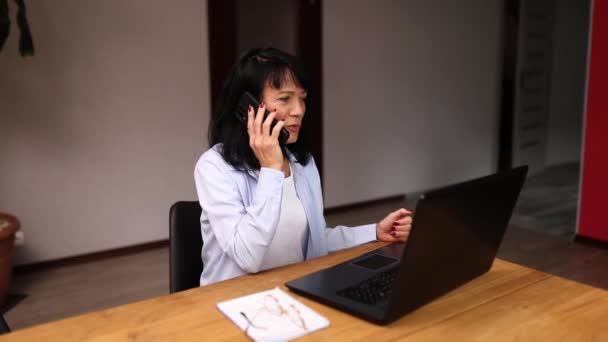 年上の女性従業員は クライアントまたはビジネスパートナーとスマートフォンコール携帯電話を持っています シニア女性はノートパソコンで職場で電話で話しています — ストック動画