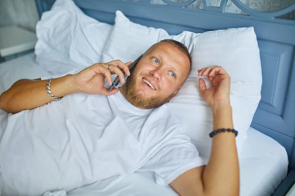 Skäggig Man Talar Smartphone Medan Liggande Mysiga Sängen Vita Sängkläder — Stockfoto