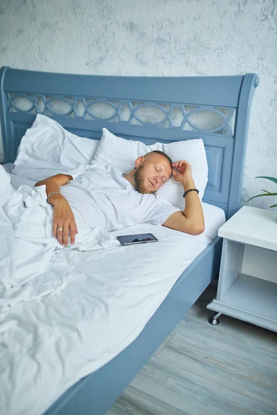 Bearded man sleeping alone on a big and cozy bed white linens with smartphone alarm at home, lifestyle, comfortable pillow, bed