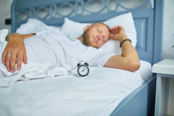 Bearded man is sleeping alone on a big and cozy bed white linens with alarm clock at home, lifestyle, comfortable pillow, bed