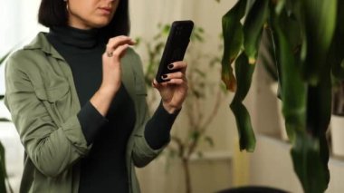 Akıllı telefonlu bir kadın evde Dracaena avuç içi yaprak hastalıklarının fotoğrafını çekiyor, ev bitkileriyle ilgileniyor, hasarlı izinler, ev bitkilerinin mobil uygulamalarında tedavi araması