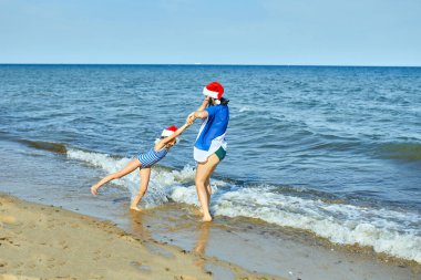 Noel Baba şapkalı mutlu anne ve kız kumsalda koşup eğleniyorlar. Yeni yıl tatilinde aile turistiyim..