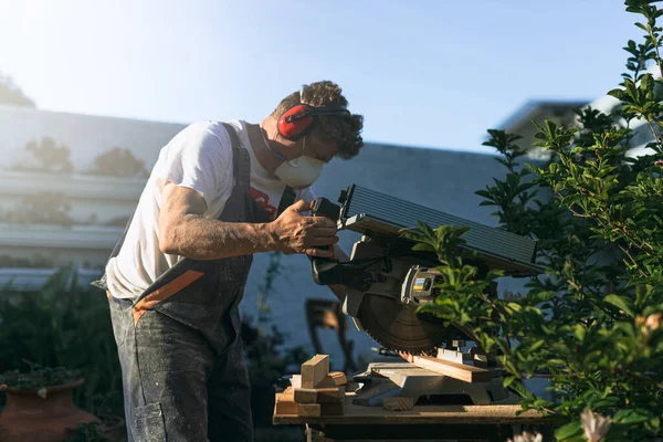 Marangoz maske takıyor ve kulaklık takıyor mavi gökyüzünde bir öğleden sonra dairesel testere ile bahçede odun kesiyor.