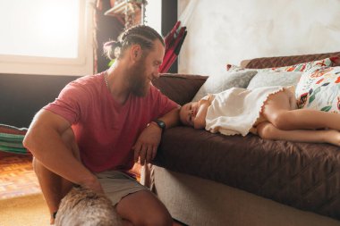 Kızıl saçlı baba kızına gülümsüyor. Evdeki kanepede yatıyor.