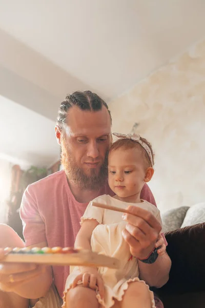 Kızıl saçlı sakallı baba evde bebeğiyle oynuyor. Oturma odasında oyuncak bir ksilofon tutuyor.