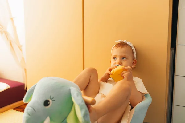 cute little girl with a toy duck in her mouth on a stuffed elephant with a yellow cupboard in the background