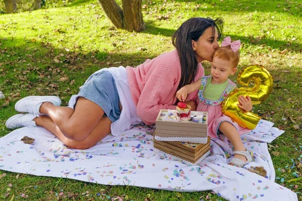 Venezuelan mother kissing her red-haired daughter during a picnic in the countryside on her second birthday eating strawberry treats under a tree