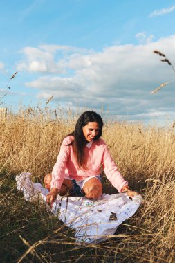 Uzun siyah saçlı, beyaz bluz giyen ve güneşli bir günde, bir buğday tarlasında battaniyeye oturmuş pembe bir kazak giyen genç bir Latin kadın.