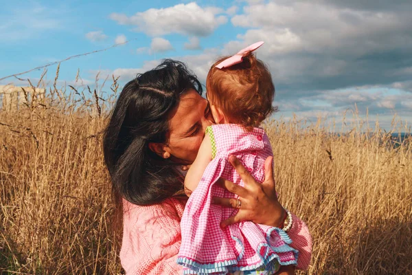 Latin siyah saçlı anne buğday tarlasında küçük kızıl saçlı kızını öpüyor ve sarılıyor.