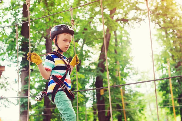 Little boy in the rope park pass obstacles. Cute kid climb the rope road. Adventure climbing high wire park. Boy wearing helmet and safety equipment. Extreme sport in adventure park.