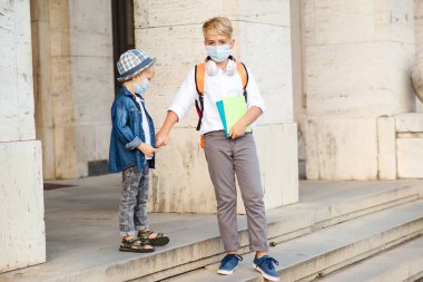 Koronavirüs salgını sırasında maske takan okul çocukları. Çocuklar okuldan sonra eve gidiyor. Coronavirus karantina ve tecrit. Tıp maskeli şirin kardeşler sokakta yürüyor..