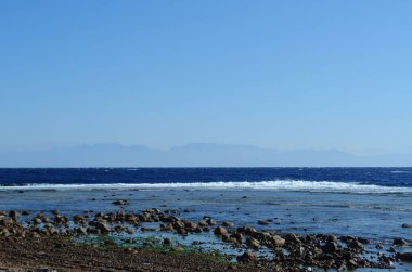 Deniz dalgaları. Deniz kıyısındaki taşlar. Diğer kıyıdaki uzak dağlar. Açık mavi gökyüzü. Mısır, Sina Yarımadası, Dahab.