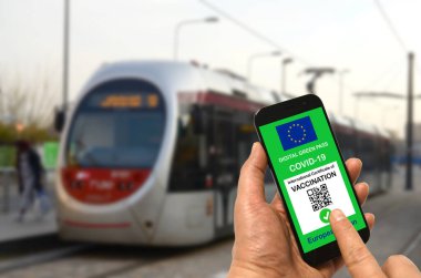 Tramvay durağındaki bir adam elinde Avrupa Birliği dijital yeşil Covid-19 kartı olan bir akıllı telefon tutuyor. Coronavirus ve Yeşil Geçiş sırasında güvenli seyahat konsepti