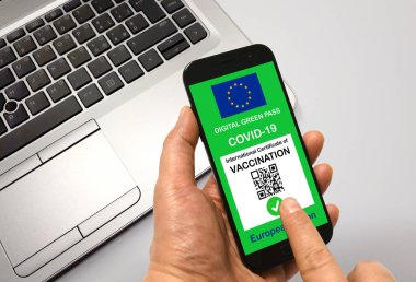 Bir adam elinde Avrupa Birliği dijital yeşil kartı olan bir akıllı telefon tutuyor. Elinde de arka planında Not Defteri olan Covid-19 için. Covid-19 için Yeşil Geçişli Mesleki Güvenlik Konsepti