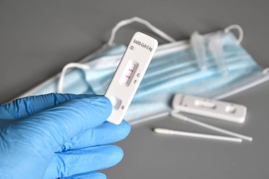 closeup of a healthcare worker, wearing blue surgical gloves, holding a covid-19 test device showing a positive result in his hand with protection mask on background