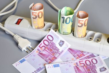 Euro banknotları yuvarlandı ve gri bir arka plandaki elektrik prizinin prizlerine yerleştirildi. Artan elektrik maliyeti kavramı. Pahalı enerji faturası ve artan elektrik fiyatları.