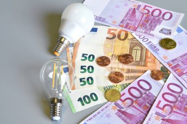 Elektrik prizi, euro para banknotları ve gri arka planda ampullü sentler. Elektrik maliyetinin artması konsepti. Pahalı enerji faturası ve elektrik fiyatlarındaki artış.