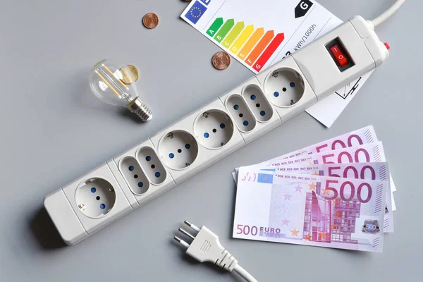 Euro banknotes and cents, light bulb, power plug of an extension socket and energy efficiency rating table on gray surface. Concept for the rising cost of electricity. Expensive energy bill and rising electricity prices.