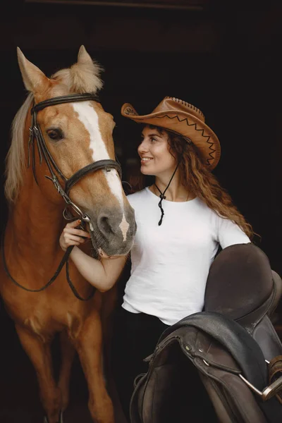 Portrait of rider woman in a leather hat with a brown horse in a stable — Photo