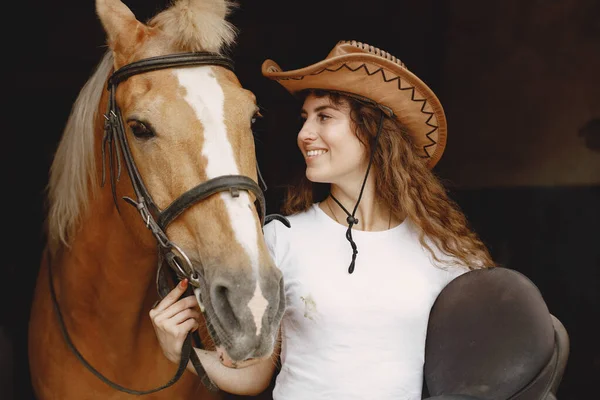 Portrait of rider woman in a leather hat with a brown horse in a stable — Photo