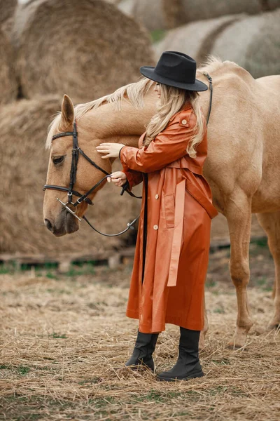 Mujer con abrigo de cuero rojo tocando un caballo marrón en una granja — Foto de Stock