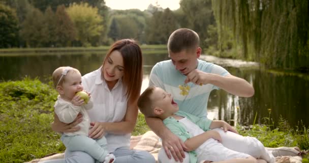 Młoda rodzina z dwójką dzieci na świeżym powietrzu nad rzeką na letnim pikniku — Wideo stockowe
