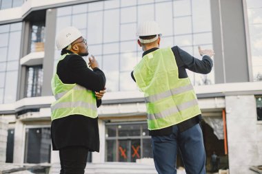 Multi racial builders standing outdoors, back view. Wearing uniform, talking about new glass building. Working on the poject. City infrastructure. Black man.