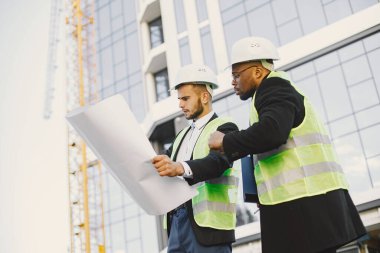 Two engineers standing near glass building. They are wearing helmets and uniform, holding blueprint and documents. Builiding, new flats, architecture.