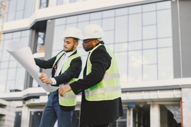 Two engineers standing near glass building. They are wearing helmets and uniform, holding blueprint and documents. Builiding, new flats, architecture.