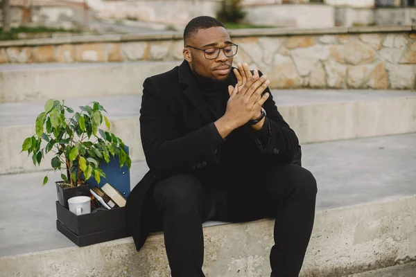 Employee get fired. Black man sitting with box on stairs and regretting.