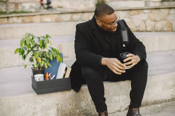 Employee get fired. Black man sitting with box on stairs and regretting.