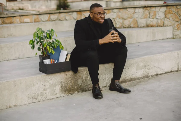 Employee get fired. Black man sitting with box on stairs and regretting.