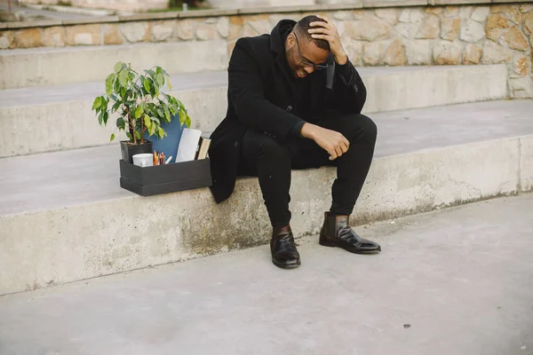 Employee get fired. Black man sitting with box on stairs and regretting.