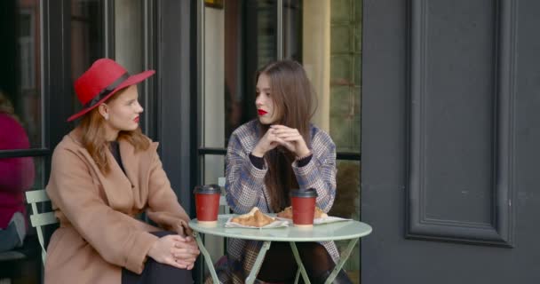 两个朋友优雅地穿着 女孩们坐在室外露台上的咖啡店里 聊天和吃早餐 — 图库视频影像