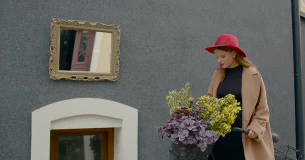 赤い帽子のかわいい女の子とバスケットに花を嗅ぐコート 彼女は町の自転車の近くに立っている — ストック動画