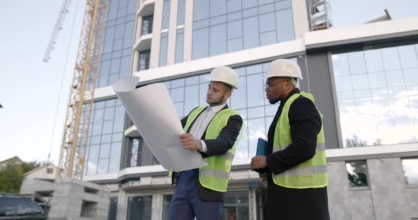 制服とヘルメットの労働者は 青写真上の新しい建物の近くの建設計画を比較します 新しい建物のコンセプト — ストック動画