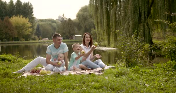 幸せな家族は 芝生のマットの上に座っている 子供と親は果物で遊ぶ 川岸の晴れ — ストック動画