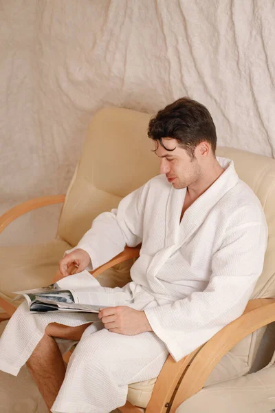 A young man is resting in the salt room. Bearded man wearing white bathrobe and reading a magazine. Spa treatments for man with respiratory disorders, inhalation.