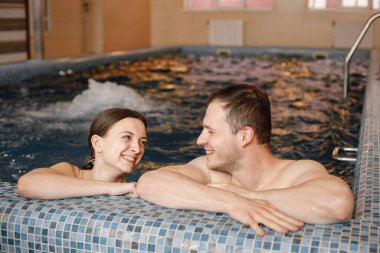 Genç çift birlikte yüzme havuzunda vakit geçiriyor. Sakallı adam ve esmer kadın havuz kenarında suyun içinde konuşuyorlar. Kadın ve erkek mayo giyiyor..