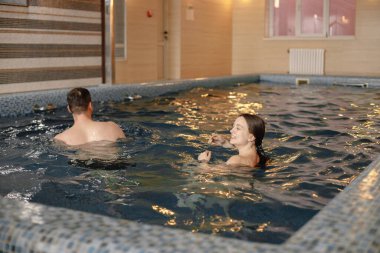Young couple is spending time together in swimming pool. Bearded man and brunette woman swimming. Man and woman wearing swimsuits.