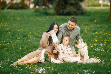 Mutlu çocuk parkta baba ve hamile anne ile oturuyor. Esmer kadın ve kızı köpekleri fransız buldoguyla çimlerin üzerinde oturuyorlar. Bej elbiseli kadın..