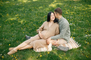 Çiftler ilk bebeklerini çimenlerin üzerinde oturup kucaklaşmayı bekliyorlar. Hamile kadın ve kocası parkta oturup fotoğraf çektirmek için poz veriyorlar. Esmer kadın bej elbise giyiyor..
